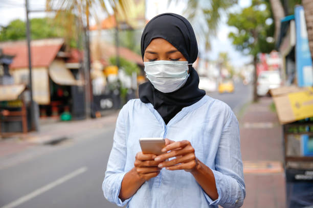 Young Muslim woman using smartphone on the street Portrait of Indonesian woman with protection face mask ,reading news about covid-19 on smartphone, while walking on sidewalk indonesian woman stock pictures, royalty-free photos & images