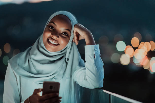 Young Muslim woman wearing scarf veil on urban city street at night texting on a smartphone with bokeh city light in the background stock photo