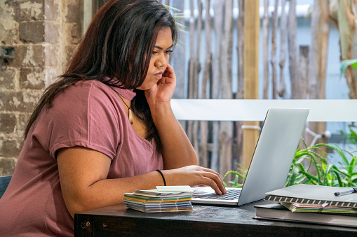This is Australia: Young  woman of Pacific Islander ethnicity looking puzzled while working on laptop computer next to sunlit window: business woman or student working from home. ID & logos edited