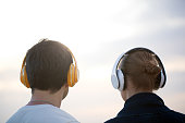 Back shot of man and woman listening to music in wireless headphones against evening cloudy sky background