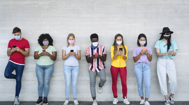 Young people wearing face mask using mobile smartphone outdoor - Multiracial friends having fun with new technology social media app during corona virus outbreak - Youth millennial lifestyle concept  generation z stock pictures, royalty-free photos & images