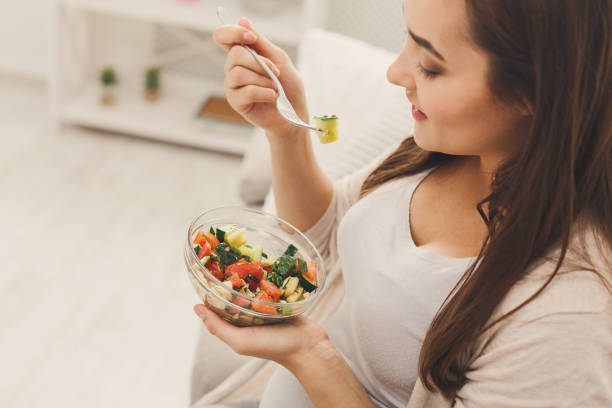 Young pregnant woman eating fresh green salad Young pregnant woman eating green salad. Attractive expectant lady sitting on sofa and having fresh snack. Healthy nutrition and pregnancy concept, copy space healthy habits during pregnancy stock pictures, royalty-free photos & images
