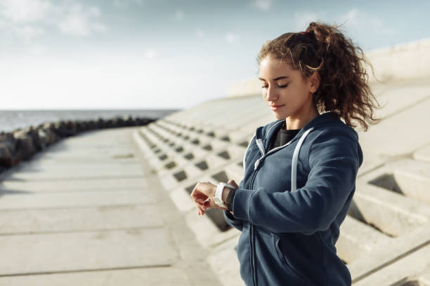 Young purposeful curly-haired fitness woman in sportswear looking at watch on city beach stock photo