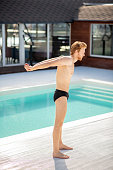 Cheerful mood. Young red-haired guy barefoot in swimming suit standing in profile, hands behind his back, by pool
