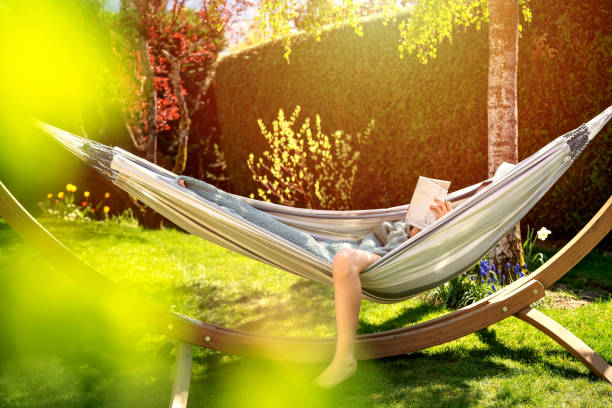 ung avslappnad flicka läser bok i hängmatta i trädgården hemma vid ljusa solnedgången. långsamt boende, gadget detox och helg fritid aktivitet. karantän- och självisoleringsperiod - hängmatta bildbanksfoton och bilder