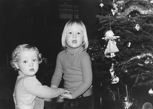 December 1977 vintage, retro monochrome image of a brother and sister, holding hands, standing next to a vintage decorated christmas tree.