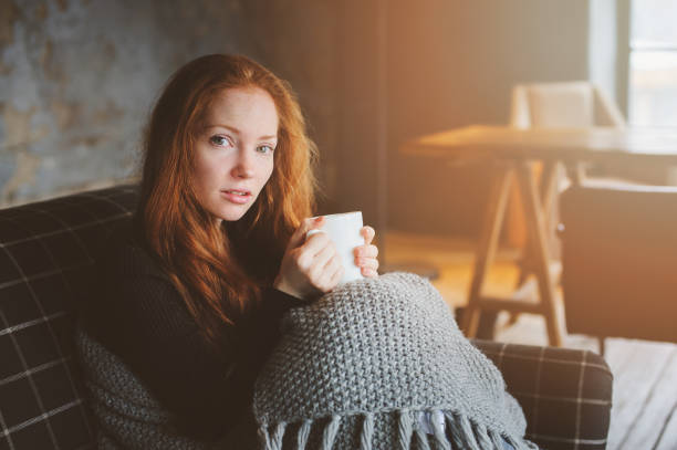 young sick woman healing with hot drink at home on cozy couch, wrapped in knitted blanket stock photo