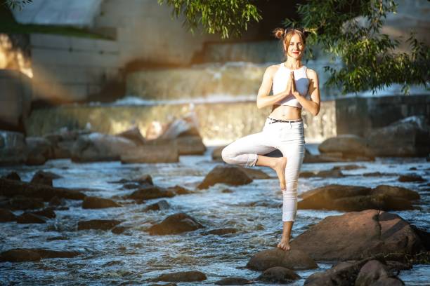 Photo of Young slender girl in white practicing yoga outdoors. Evening meditation and relaxation at the river and waterfall. The Tree position of yoga (Vrikshasana).