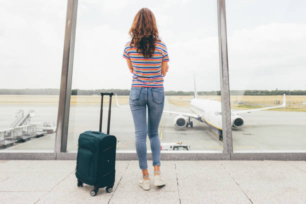 Young slim woman waiting her flight in lounge hall (just after duty free) in airport with her hand luggage (green trolley). Lost or canceled flight, need compensation concept. Eindhoven, Netherlands stock photo
