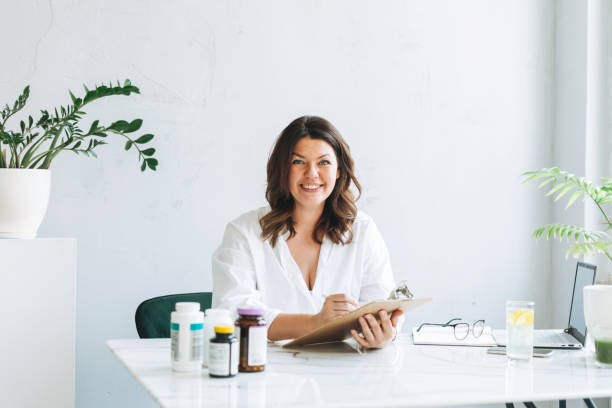 Young smiling brunette woman doctor nutritionist plus size in white shirt working at laptop at modern bright office room. The doctor prescribes a prescription for medicines and vitamins at clinic stock photo