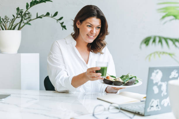 Young smiling brunette woman nutritionist doctor plus size in white shirt with green healthy food at modern bright office room. Doctor communicates with patient online stock photo
