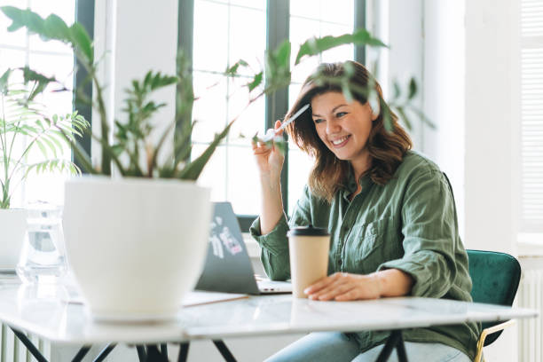 Young smiling brunette woman plus size working at laptop on table with house plant in the bright modern office stock photo