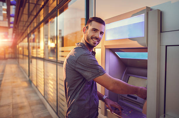 Young smiling businessman withdrawing money from ATM. Young happy businessman withdrawing money from a cash machine and looking at the camera. banks and atms stock pictures, royalty-free photos & images
