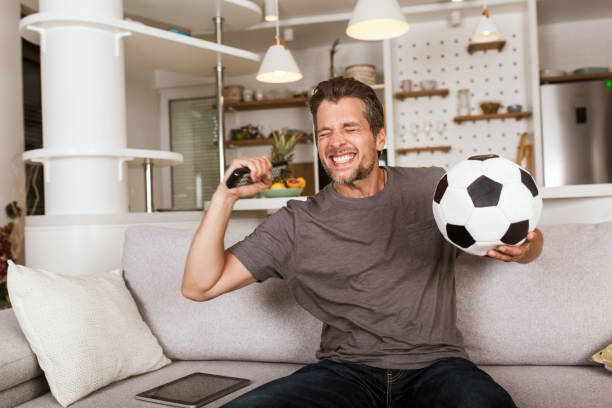 Young soccer fan man watching football game on television at living room couch. stock photo