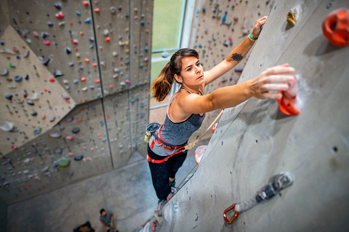 High level close-up of Caucasian woman in early 20s reaching for hand hold on wall in modern sport climbing gym.