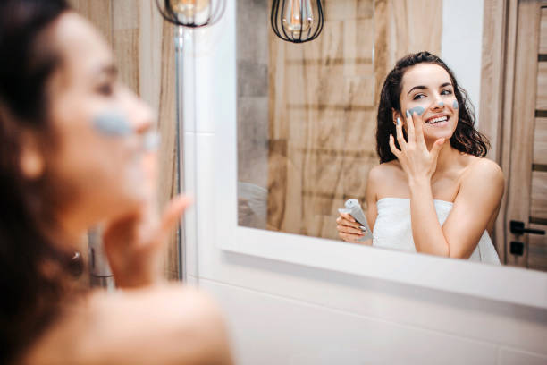 Young sporty dark-haired beautiful woman doing morning evening routine at mirror. Cheerful happy model wrapped in white towel and put mask on face. stock photo