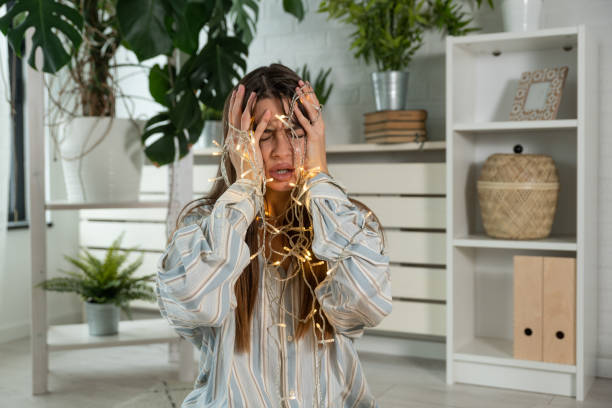 Young stressed and frustrated woman sitting at home having issue with tangled Christmas wire lights. Female with problems of untie decoration for Holidays. stock photo
