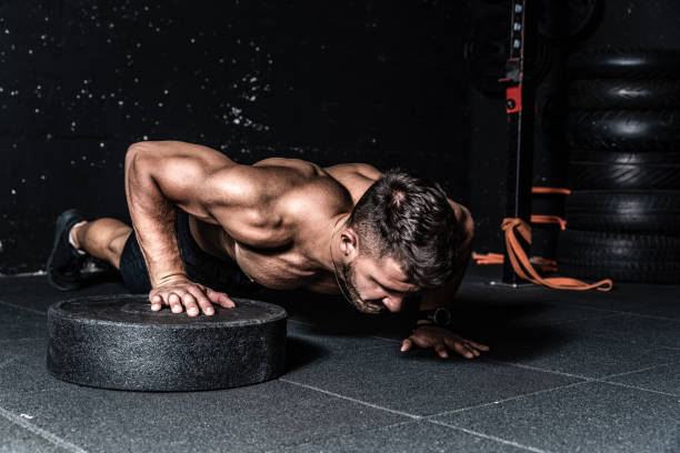 Young strong sweaty focused fit muscular man with big muscles doing push ups with one hand on the barbell weight plate for training hard core workout in the gym real people selective focus stock photo