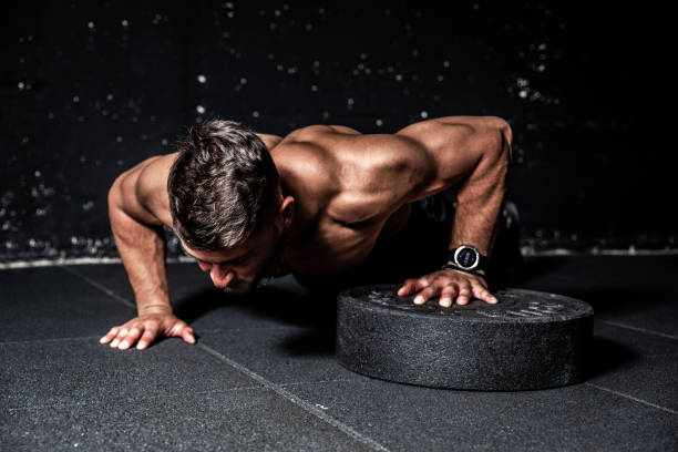 Young strong sweaty focused fit muscular man with big muscles performing push ups with one hand on the barbell weight plate for training hard core workout in the gym stock photo