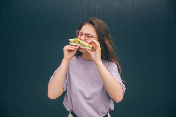 Young stylish trendy woman isolated over grey blue background. Hungry girl biting piece of fresh sandwich. Woman having breakfast or lunch outside at wall. Eating alone. Young stylish trendy woman isolated over grey blue background. Hungry girl biting piece of fresh sandwich. Woman having breakfast or lunch outside at wall. Eating alone vlad model photos stock pictures, royalty-free photos & images