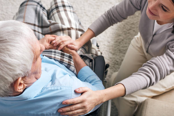 Young supportive female caregiver sitting by senior man in wheelchair Young supportive female caregiver sitting by senior man in wheelchair and keeping her hand on his shoulder while comforting him assisted living stock pictures, royalty-free photos & images
