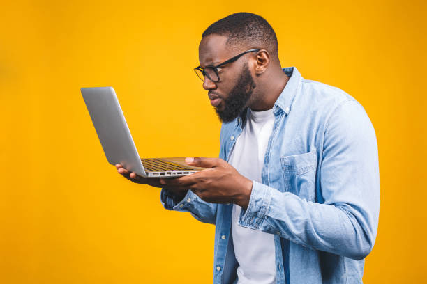 Young surprised african american man standing and using laptop computer isolated over yellow background. Young surprised african american man standing and using laptop computer isolated over yellow background. worried man funny stock pictures, royalty-free photos & images