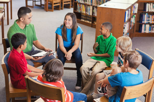 Young teacher leads after school club in library In this view from above, a young male teacher sits in a circle of chairs with a group of students after school.  They are having a club meeting in the school library. middle school teacher stock pictures, royalty-free photos & images