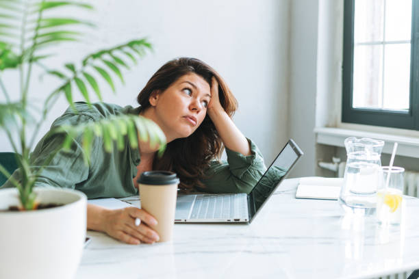 Young thinking unhappy brunette woman plus size working at laptop on table with house plant in the bright modern office stock photo