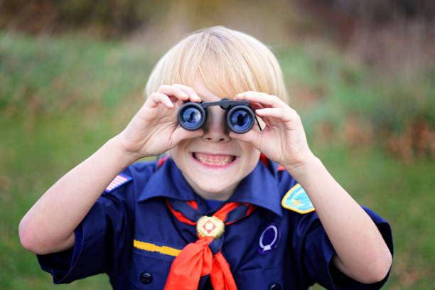 Young Tiger Cub Scout Smiling at Camera Through Binoculars A young Cub Scout in the tiger den is smiling as he looks at the camera through small binoculars outside on a summer day. scout camp stock pictures, royalty-free photos & images