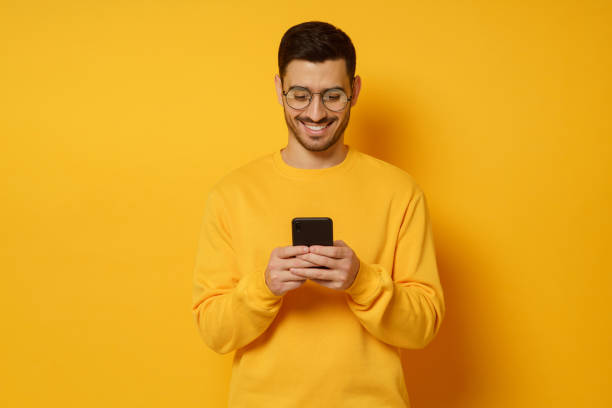 jonge trendy mens in glazen en sweater, die gelukkig over gesprek in tekstberichten met vrienden via telefoon voelen, glimlachend terwijl het bekijken van scherm, dat op gele achtergrond wordt geïsoleerd - gekleurde achtergrond stockfoto's en -beelden