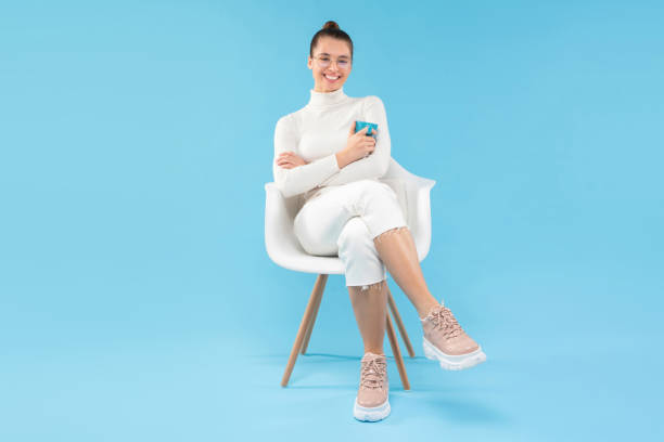joven chica positiva de moda sentada en el sillón, sosteniendo la taza de café en las manos, disfrutando de su tiempo libre del trabajo, aislada sobre fondo azul - sentado fotografías e imágenes de stock