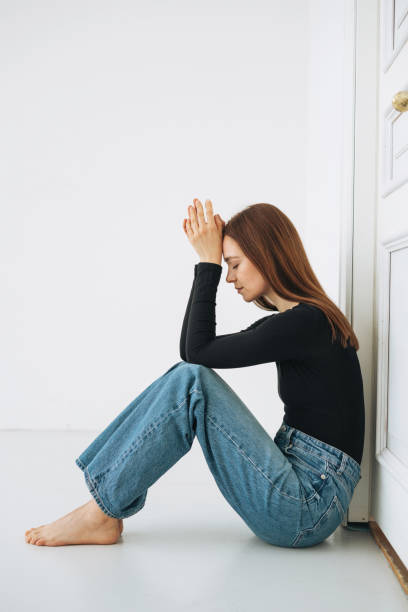 Young unhappy woman teenager girl with long hair in jeans sitting on floor in closed position by the door at home, negative emotion stock photo