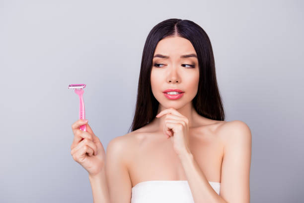 Young unsure nervous asian girl with pink shaver is standing isolated on a grey background, wrapped in a white towel, doubting if she should use it Young unsure nervous asian girl with pink shaver is standing isolated on a grey background, wrapped in a white towel, doubting if she should use it women shaving stock pictures, royalty-free photos & images