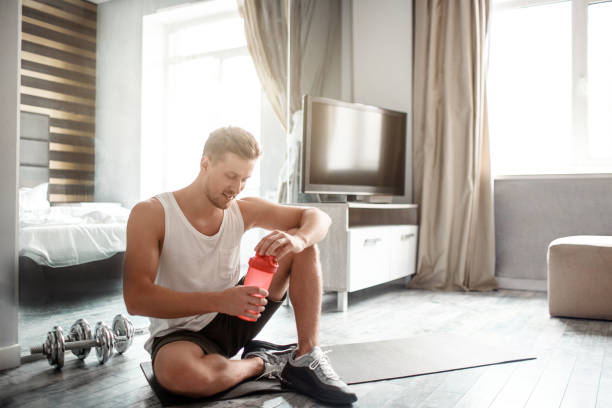 Young well-built man go in for sports in apartment. Nice guy sit on carimate and hold red bottle with water. Workout break. Smile. stock photo