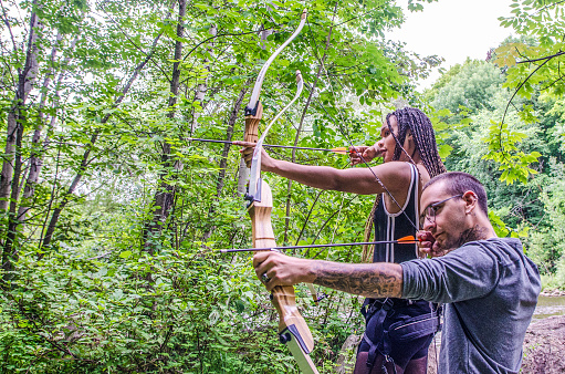 Young white man and Haitian woman using bow in forest during day of summer