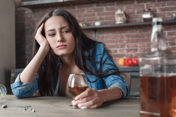 Young woman alcoholic social problems concept sitting looking at bottle of whiskey Young female alcoholic social problems sitting at table drinking whiskey looking drunk at bottle close-up alcohol abuse stock pictures, royalty-free photos & images