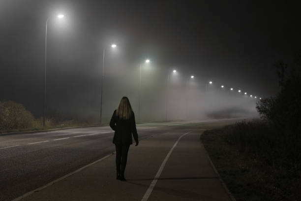 young woman alone slowly walking under white street lights in night. dark time. peaceful atmosphere in mist. foggy air. back view. - trilhos pedestres imagens e fotografias de stock