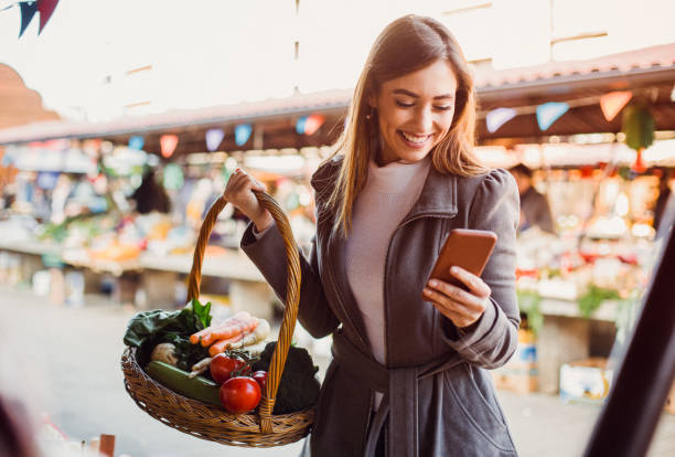 en ung kvinna på den gröna marknaden shopping för grönsaker, titta på inköpslistan på hennes telefon. - squash sun bildbanksfoton och bilder