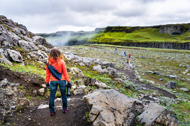 Young woman back standing looking at Dettifoss waterfall on rocks water mist spraying cloudy day in Iceland with orange jacket and jeans Young woman back standing looking at Dettifoss waterfall on rocks water mist spraying cloudy day in Iceland with orange jacket and jeans iceland dettifoss stock pictures, royalty-free photos & images