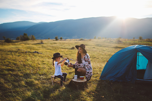 Landscape in Dinaric Alps, mountain range in Southern Europe, on the border of Croatia and Bosnia. Young brunette woman is camping with her baby girl child. They are sitting in the in front of a small tent, enjoying a beautiful view over the valley under the mountains, watching the horizon as the sun sets, playing games and having fun in the nature outdoors. They're dressed in fashionable hippie clothes, enjoying freedom in the nature, wearing hats and having a coffee in a small stove top espresso pot.