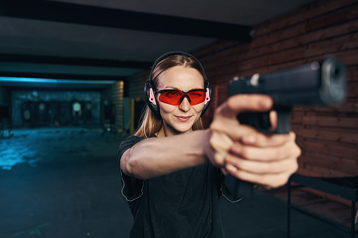 Waist-up portrait of a focused female sniper in safety glasses and earmuffs firing a pistol with both hands
