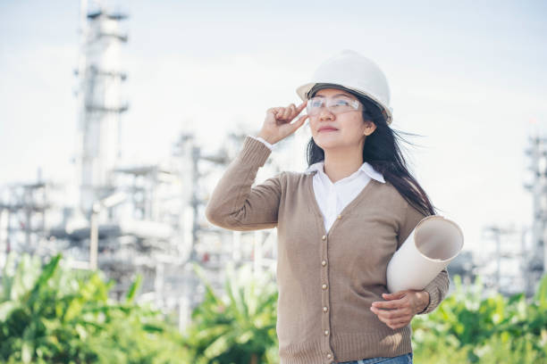 Young woman engineer or construction worker wearing safety hat (helmet) and holding blueprint with refinery plant background. stock photo