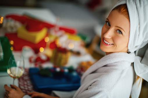 Young woman wearing a bathrobe and wrapped towel on her head, drinking white wine and preparing to decorate Christmas presents. She's smiling and looking at camera.