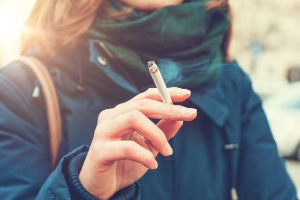 Young woman enjoying a cigarette Young woman enjoying a cigarette outdoors holding it between her fingers, low angle view against the chest of a warm autumn jacket in a smoking and tobacco concept smoking activity stock pictures, royalty-free photos & images