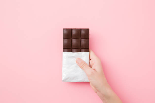 Young woman hand holding chocolate bar on light pink table background. Opened pack. Sweet snack. Closeup. Top down view. Young woman hand holding chocolate bar on light pink table background. Opened pack. Sweet snack. Closeup. Top down view. dark chocolate stock pictures, royalty-free photos & images