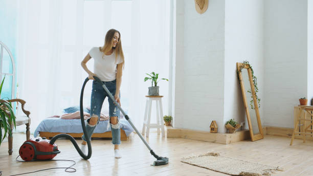Young woman having fun cleaning house with vacuum cleaner dancing and singing at home stock photo
