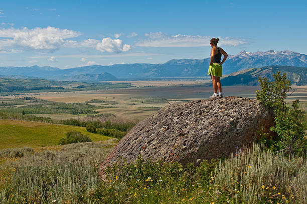 Young Woman Hiker Standing on Coyote Rock and Overlooking the Jackson Hole Valley Across the valley from the Tetons is the Gros Ventre range. In Wyoming they say people come to visit the Tetons and end up falling in love with the Gros Ventres. What these mountains lack in height and rugged grandeur they more than make up for with their gentle beauty and sweeping vistas. Their brightly colored alpine meadows are a joy of sights and smells. This young woman hiker was photographed standing on Coyote Rock in Bridger Teton National Forest near Kelly, Wyoming, USA. jeff goulden grand teton national park stock pictures, royalty-free photos & images