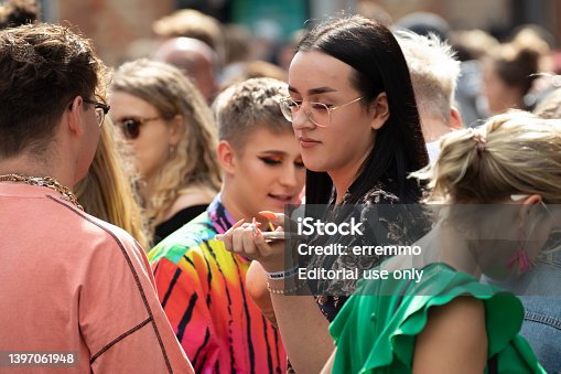 Young woman hold mobile phone in crowd with a male in rainbow colours in the background.