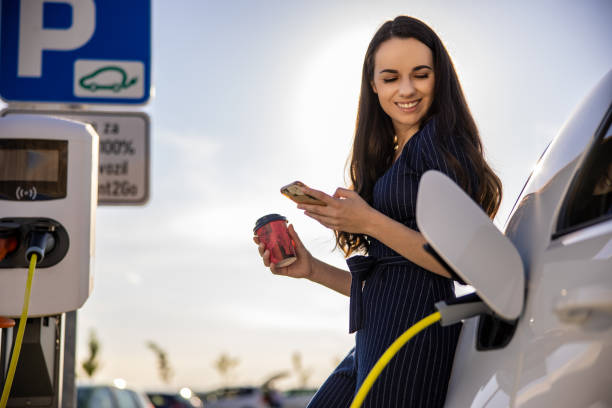 young woman holding mobile phone and cup while waiting for electric car to charge - electric car woman bildbanksfoton och bilder