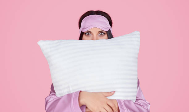 Young woman hugging soft pillow Young female in silk lilac sleep mask and pajama looking at camera and embracing comfortable pillow during bedtime against pink background eye mask stock pictures, royalty-free photos & images